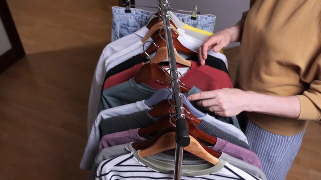 Person's hands selecting a t shirt on a crowded clothing rack, organizing a home wardrobe with wooden and plastic hangers for a neat, everyday closet and outfit selection routine