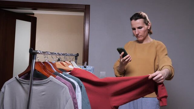 Woman photographing a sweatshirt on a clothing rack with a smartphone, preparing second hand fashion items for online resale and social media listing from home studio