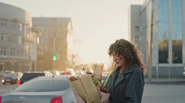 Mobile lady with blossoms present. City lady pauses at crossroads with vibrant flower bouquet in hand. Woman in metropolis pauses at intersection holding lively collection of floral stems