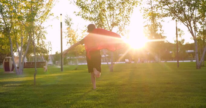 Superhero child runs through park at sunset light. Superhero cape flows behind in golden sun. Kid boy imagines adventure and hero journey outdoors. Creativity, dream, inspiration in childhood. Happy