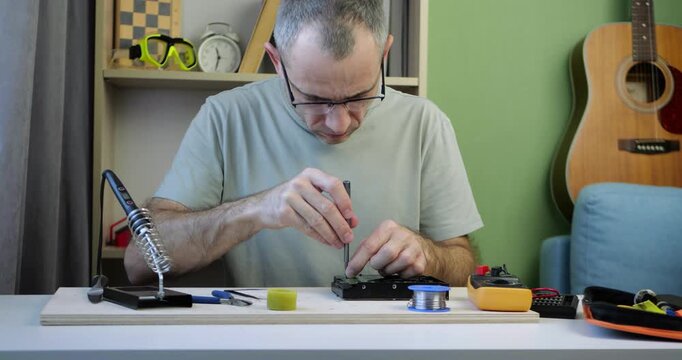 Man repairs electronic device on table. Tools and components surround technician. Focused work showcases technical skills. Ideal for technology, lifestyle, and DIY projects.