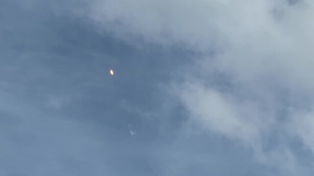 A rocket flies high in the sky with a bright glowing flame visible against soft clouds. The spacecraft appears distant as it continues its ascent through the upper atmosphere.
