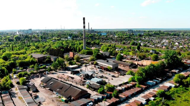 Aerial drone view of an industrial zone in Sumy, Ukraine featuring railway tracks, factory buildings, and a tall smokestack rising above the landscape. The scene includes rows of metal garages