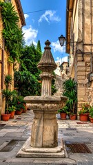Fototapeta premium A stone fountain sits in a narrow cobbled street, adorned with potted plants and surrounded by tall buildings under a blue sky