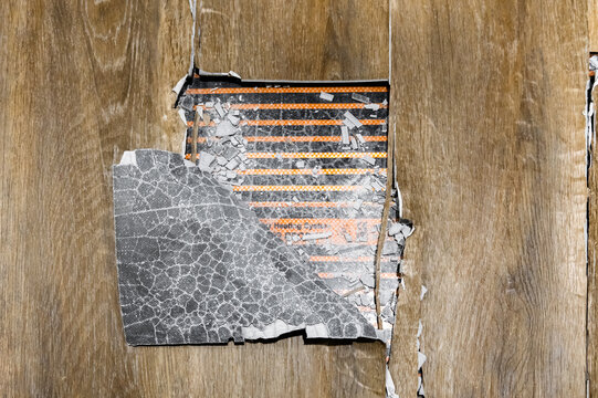 closeup torn flooring revealing foil heating mat. cracked foil patch and exposed thermal element. textured laminate surface with broken backing. diagnostic inspection