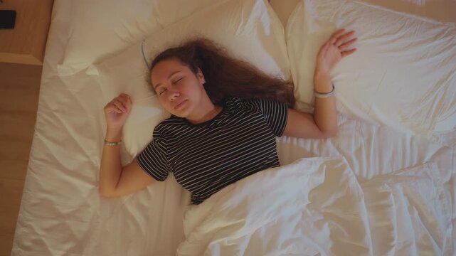 Nervous woman resting. Woman appears restless on hotel bed. Unsettled woman lying in dimly lit hotel room. Candid overhead image of woman experiencing discomfort in hotel bed under soft lighting