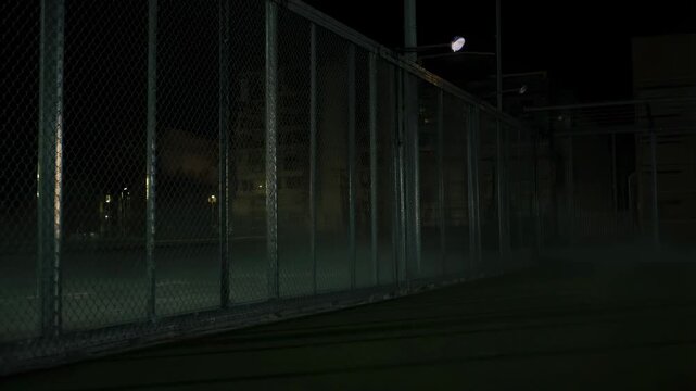 Panning tracking shot of chain link fence sliding to reveal illuminated artificial turf sports field at night