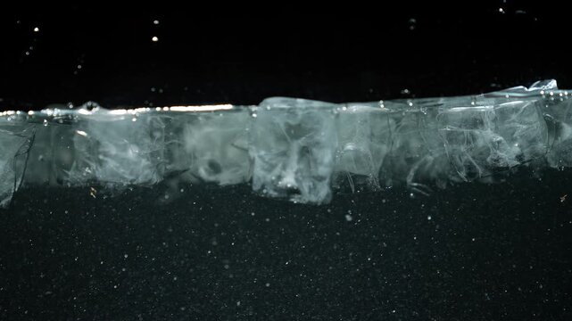 Close up of ice layer floating on water with oxygen bubbles and particles in dark background. Concept of cold temperature, purity, refreshment, science, beverage, climate, and freezing process.