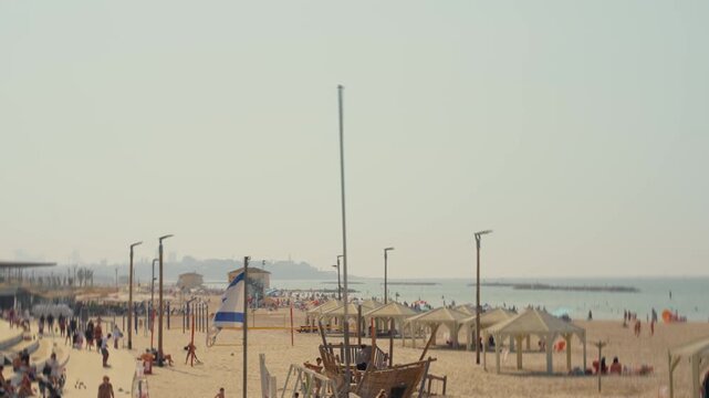 Israel flag waving by Tel Aviv beach promenade and coast on sunny summer day. Weekend holiday or summer vacation. People enjoy hot weather bathing swimming in sea or running along beautiful coast