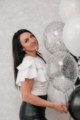 Naklejka premium A young Caucasian woman with long dark hair poses with a cluster of balloons in black, white, and silver. The background is light and neutral.