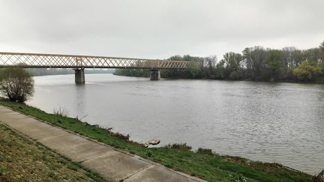 Wide view of Tisa river with bridge and embankment under cloudy sky in Senta Serbia. Concept of calm nature, travel, and peaceful atmosphere