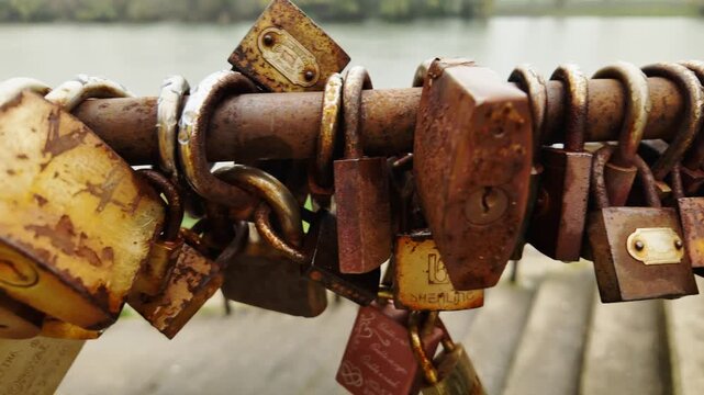 Multiple rusty love padlocks attached to metal railing near river, symbolizing relationships, commitment and romantic tradition