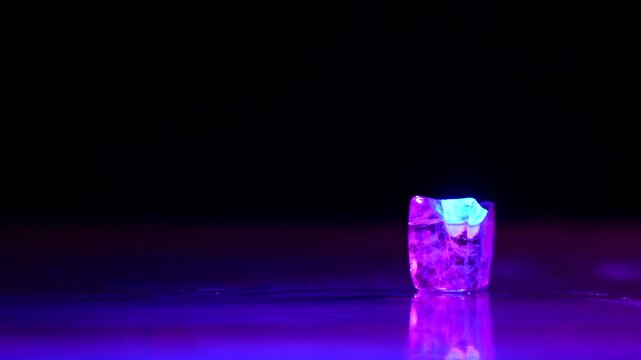 Single ice cube glowing in purple and blue light on a reflective dark surface with copy space. Concepts for advertising, nightlife, drinks in bars and restaurants, and a refreshing nightclub