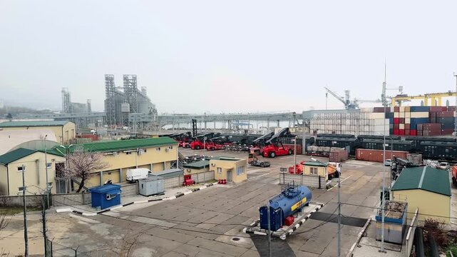 Authentic wide panorama panning across Novorossiysk commercial sea port industrial terminal with red reach stackers freight train wagons container stacks and massive grey grain silo complex