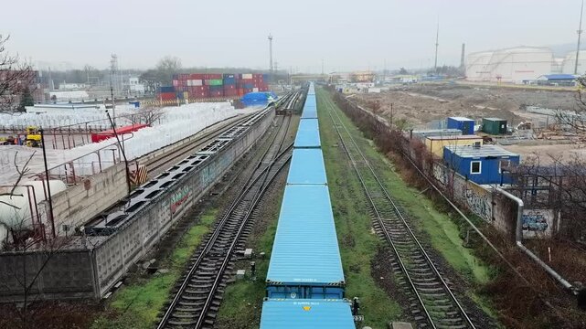 High angle view from bridge of long freight train transporting blue and red shipping containers on railroad tracks through industrial district in Novorossiysk during overcast day perspective shot
