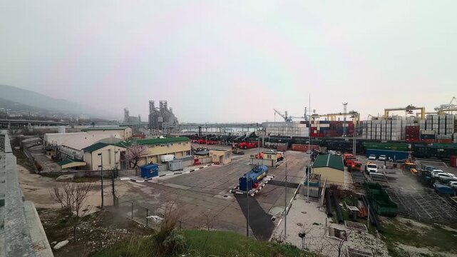 Ultra wide angle panorama of Novorossiysk sea port industrial terminal logistics hub with red reach stackers freight train wagons and massive grain silos under overcast sky authentic wide shot