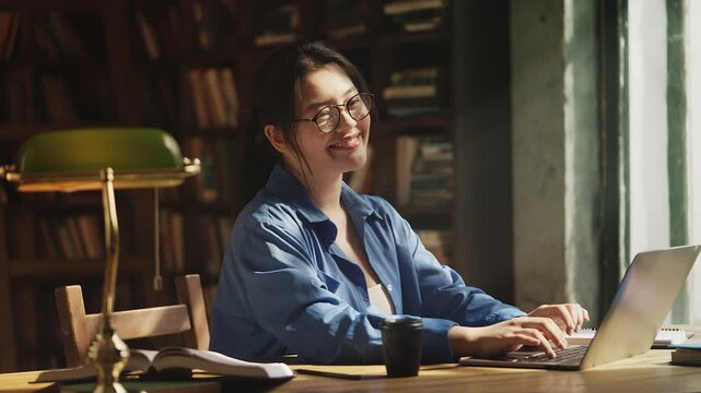 Happy Asian female student typing on laptop in university library, education