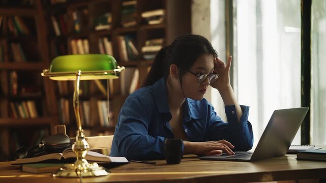 Exhausted Asian female student typing on laptop in a library, work overload