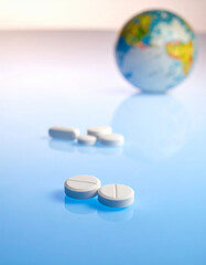 Pair of pills in foreground with blurred world globe and medication on light blue background.