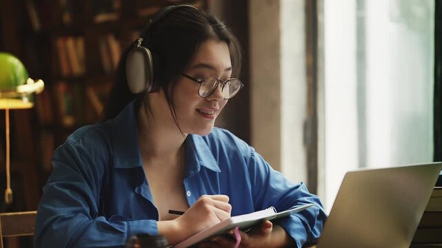 Asian female student in headset having video lesson on laptop, taking notes