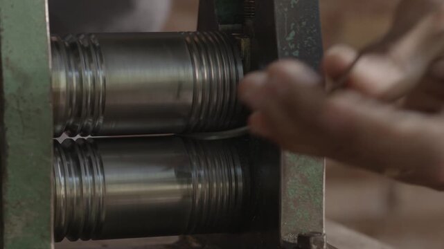 Hands guide metal wire through rolling machine. Tool applies pressure to shape material with precision. Workshop setting suggests skilled craftsmanship and manual labor