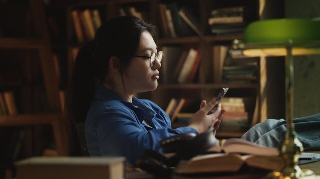 Bored Korean woman scrolling social media on phone, procrastinating in a library