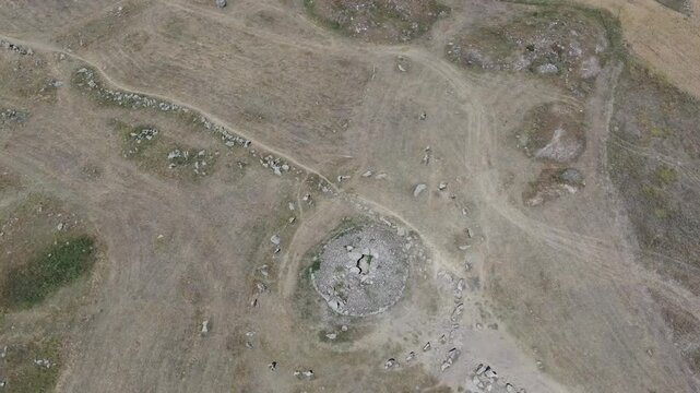 Aerial drone view flying over an ancient historical megalithic monument featuring a circle of standing stones and a central burial mound set in a dry, grassy rural landscape.