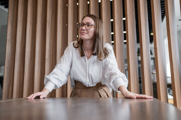 Fototapeta premium Young woman with glasses smiling at desk in modern office with wood panel wall. Smiling, glasses, desk, office, young woman, panel wall.