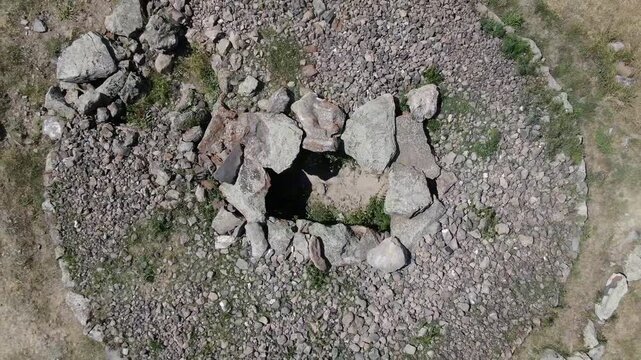 Aerial drone view flying over an ancient historical megalithic monument featuring a circle of standing stones and a central burial mound set in a dry, grassy rural landscape.