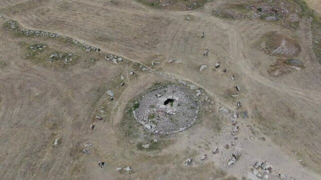 Aerial drone view flying over an ancient historical megalithic monument featuring a circle of standing stones and a central burial mound set in a dry, grassy rural landscape.