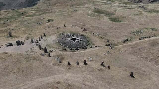 Aerial drone view flying over an ancient historical megalithic monument featuring a circle of standing stones and a central burial mound set in a dry, grassy rural landscape.