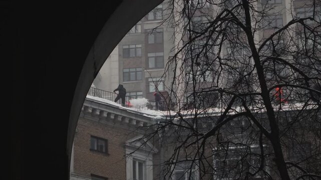 Rope access snow shoveling from roof of building. Man uses a shovel to clean a house during a blizzard. Snowfall in winter in Russia. Male worker janitor throws or drop ice, frost. Apartment building