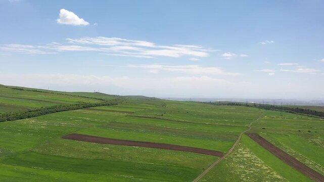 Aerial panning shot of a lush green mountain slope covered in agricultural fields. The bright blue summer sky features scattered fluffy white clouds above the rolling hills landscape.