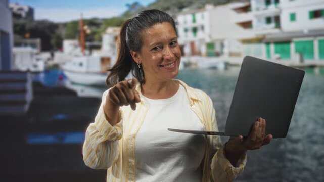 Woman hispanic middle age brunette holding laptop in left hand and pointing index finger toward camera while smiling in studio seascape set; confidence friendly teaching.