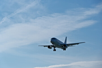 Obraz premium White passenger plane on final approach on a sunny day with blue sky, light clouds and beautiful backlighting. Sunlit white airplane landing against a blue sky with wispy clouds, backlit by bright sun