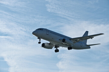 Fototapeta premium White passenger plane on final approach on a sunny day with blue sky, light clouds and beautiful backlighting. Sunlit white airplane landing against a blue sky with wispy clouds, backlit by bright sun