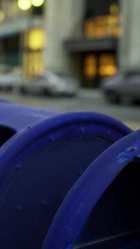 A blue mailbox stands on a city street. Cars are parked nearby. Buildings with lights are visible in the background. It is late afternoon and the street is busy.