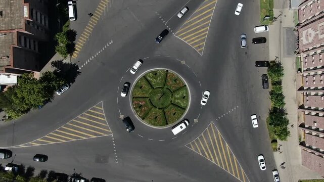 Overhead aerial view of a busy city traffic roundabout. Cars and a bus navigate the circular intersection featuring a green center, surrounded by urban buildings, trees, and sidewalks.