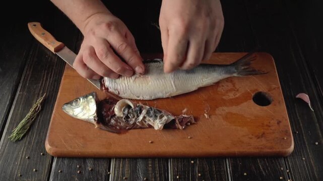 Hands of a person filleting a herring fish on a wooden cutting board, with a knife and fresh herbs visible in a rustic kitchen setting
