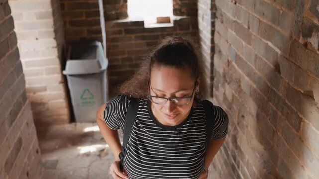 Young woman exploring brick tower interior, touching rough masonry, peering through narrow windows, carrying backpack and wearing glasses, warm sunlight casting shadows across stone corridors,