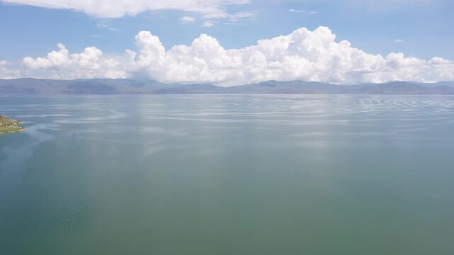  Aerial drone footage flying over a vast, calm lake with unique surface ripples, heading towards majestic distant mountains under a bright blue sky filled with large fluffy clouds.