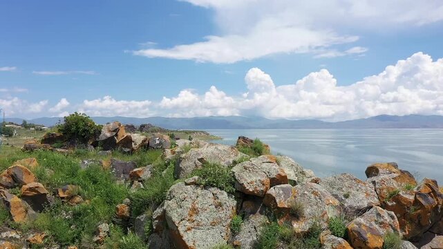 A drone flies backward over a rocky, grassy hill to reveal a beautiful ancient stone monastery complex overlooking a vast, calm blue lake and distant majestic mountain landscape.