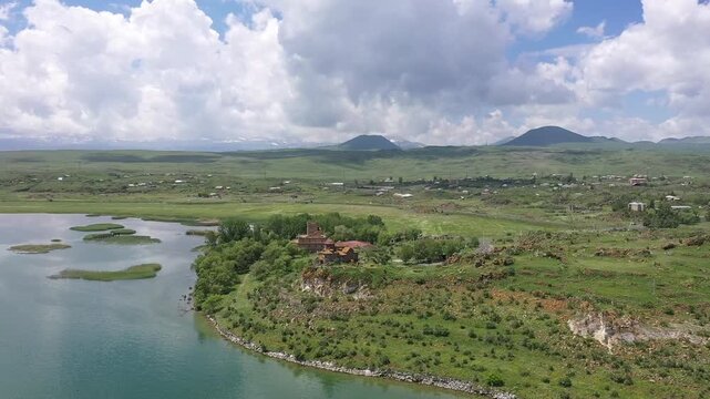 A drone flies backward over a rocky, grassy hill to reveal a beautiful ancient stone monastery complex overlooking a vast, calm blue lake and distant majestic mountain landscape.