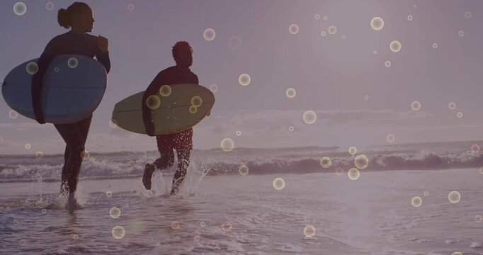 Couple spotting waves and running into surfing with boards while bubble bokeh drifting across frame