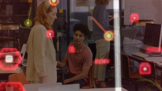 Seated man looking as leaning peer approaching in business, red hexes over monitors mapping project