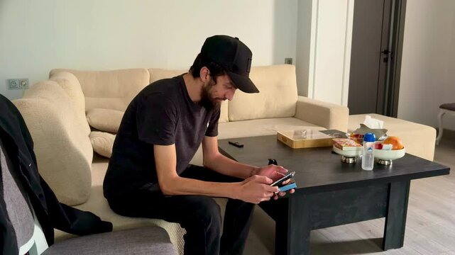 Man in Casual Clothes and Baseball Cap Sitting on Sofa in Modern Minimalist Interior, Focused on Using Smartphone and Browsing Social Media or Texting against Coffee Table