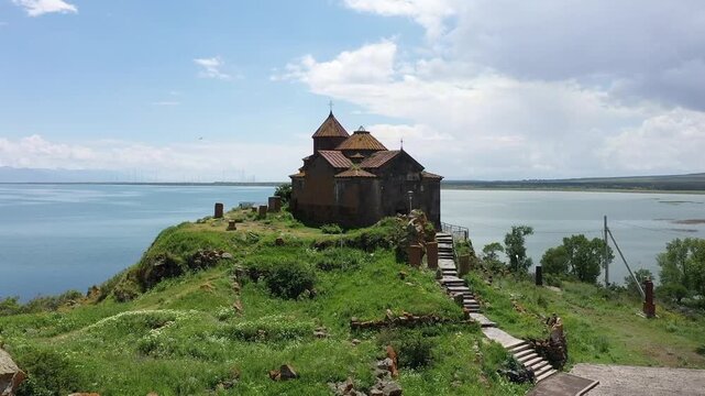 A drone flies backward over a rocky, grassy hill to reveal a beautiful ancient stone monastery complex overlooking a vast, calm blue lake and distant majestic mountain landscape.