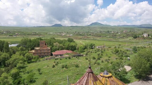 A drone flies backward over a rocky, grassy hill to reveal a beautiful ancient stone monastery complex overlooking a vast, calm blue lake and distant majestic mountain landscape.