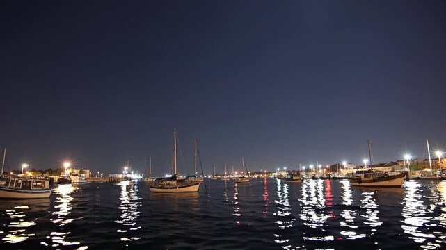 Night harbor with moored boats reflecting city lights, calm rippling water and illuminated piers under deep indigo sky serene marina atmosphere with gentle wave motion, distant skyline glow, ideal