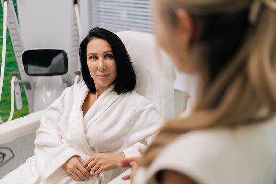 Attractive middle-aged woman client in bathrobe attentively listening to cosmetologist during in-clinic consultation about personalized aesthetic treatments, skin care plans and wellness options.